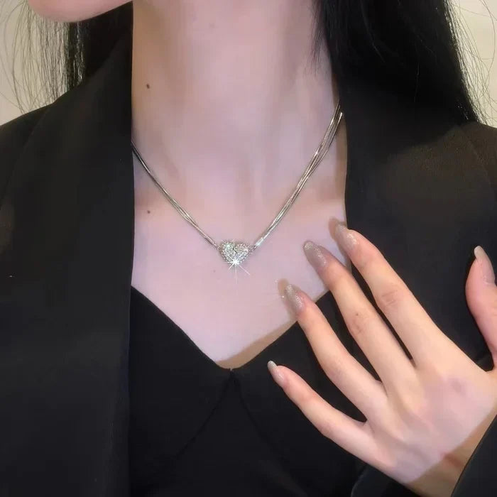 Close-up of a woman in a black blazer wearing the layered silver chain with a sparkling rhinestone heart, one hand resting near her collar. 2