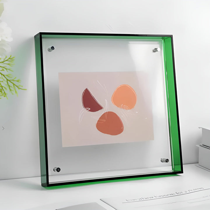 Square clear acrylic frame with a green edge and four metal standoff screws, holding an abstract print and standing on a white desk. 21