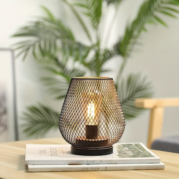 One lit mesh lantern on a stack of two magazines on a wooden table, with a blurred palm plant behind. 10