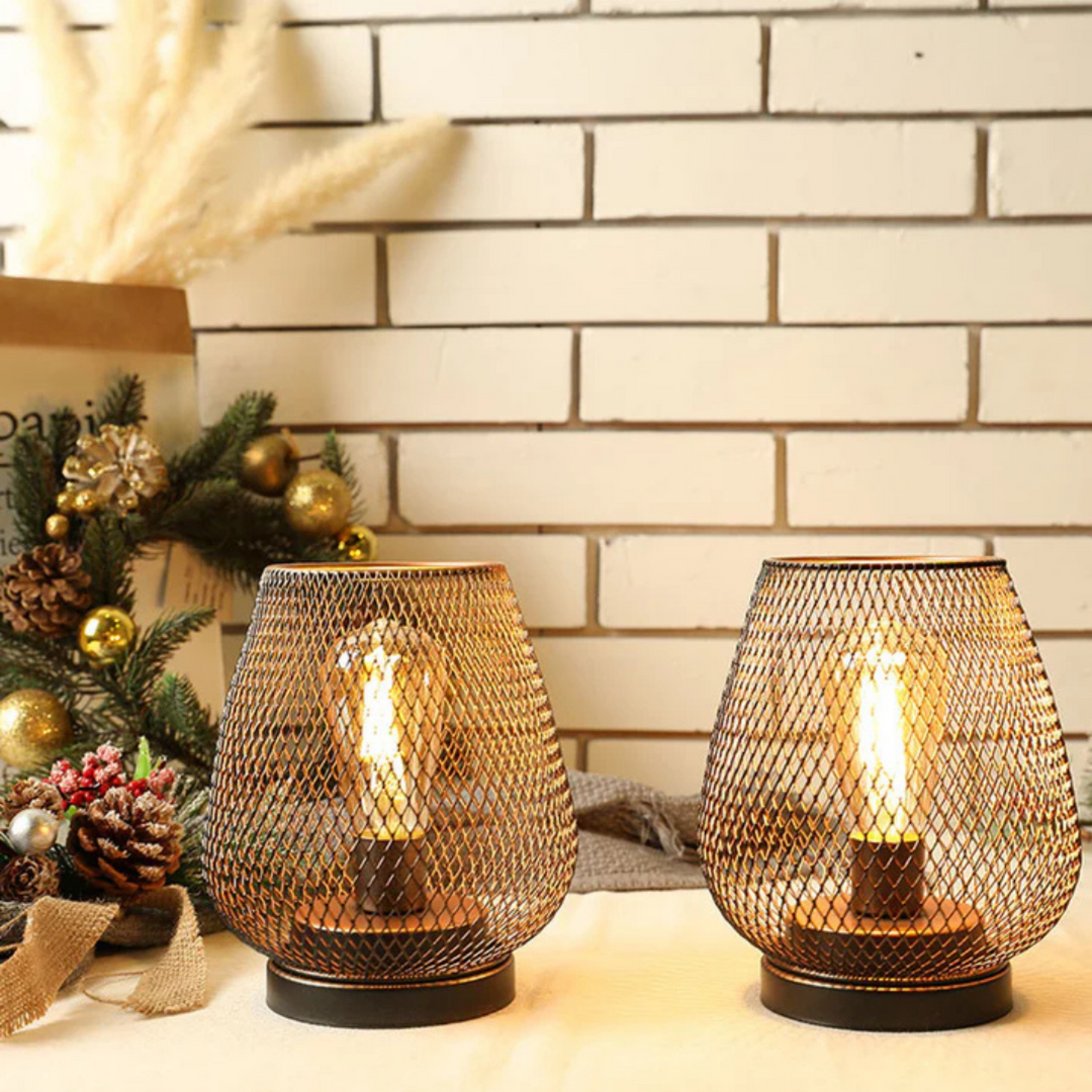 Two lit mesh lanterns on a table in front of a white brick wall, next to pine branches, pinecones and gold ornaments. 7