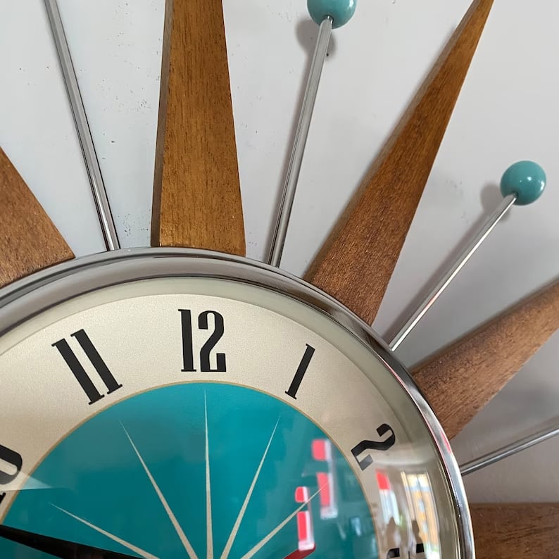 Close-up detail of the top of the starburst wall clock, showing tapered wooden spikes, chrome rods, teal balls and the domed dial glass.