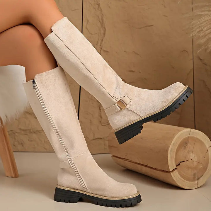 Beige knee-high boots with the side zipper, buckle strap and black treaded sole.