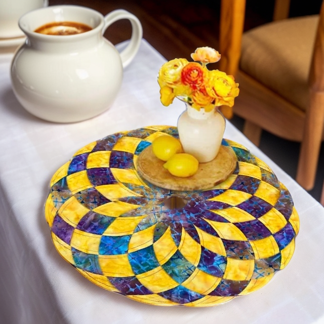 Round pieced placemat in yellow, purple and teal holding a small vase of orange flowers and lemons.