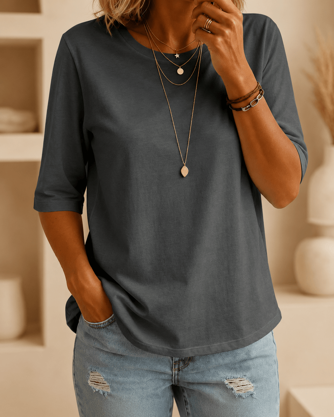 Woman wearing a dark gray half-sleeve crew-neck tee with layered gold necklaces and light distressed jeans, one hand in her pocket. 1