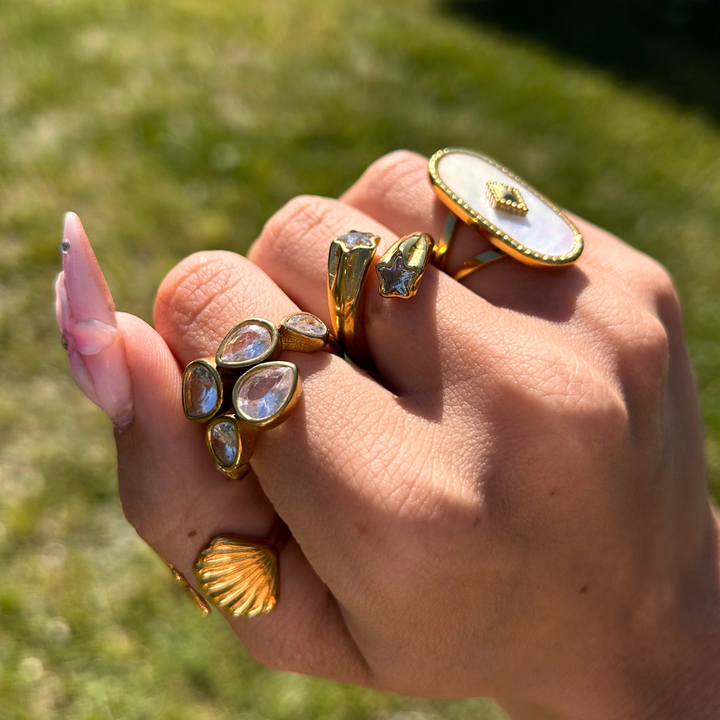 Closed hand outdoors on grass wearing the gold star ring alongside teardrop crystal, pearly oval and shell-shaped rings. 4