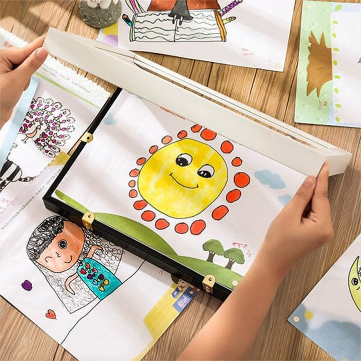Child's hands lifting the front panel of a kids art frame on a table, with a smiling sun drawing and other artwork around it.