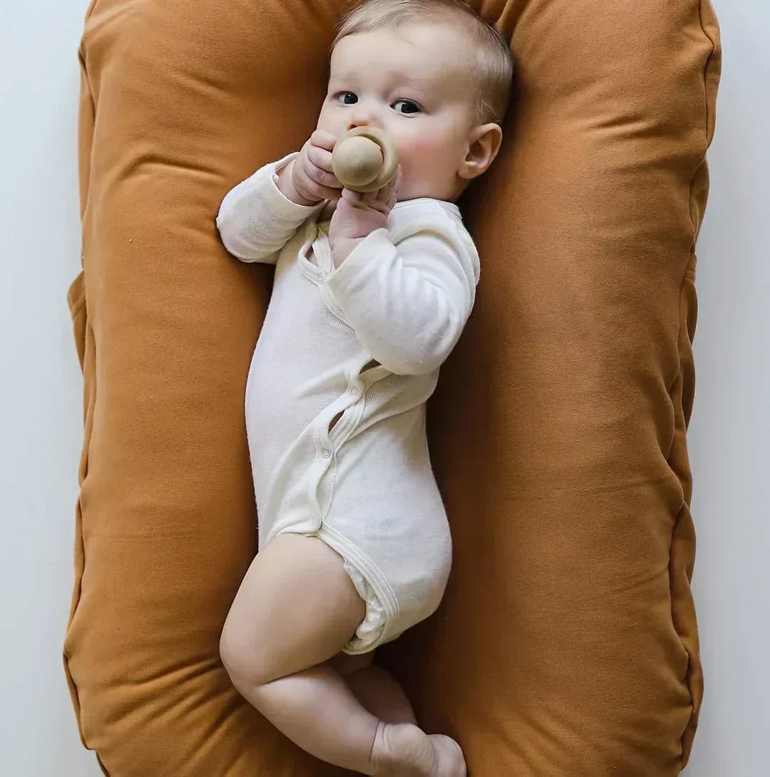 Baby in a cream bodysuit holds a wooden toy while lying in a caramel-colored lounger 4