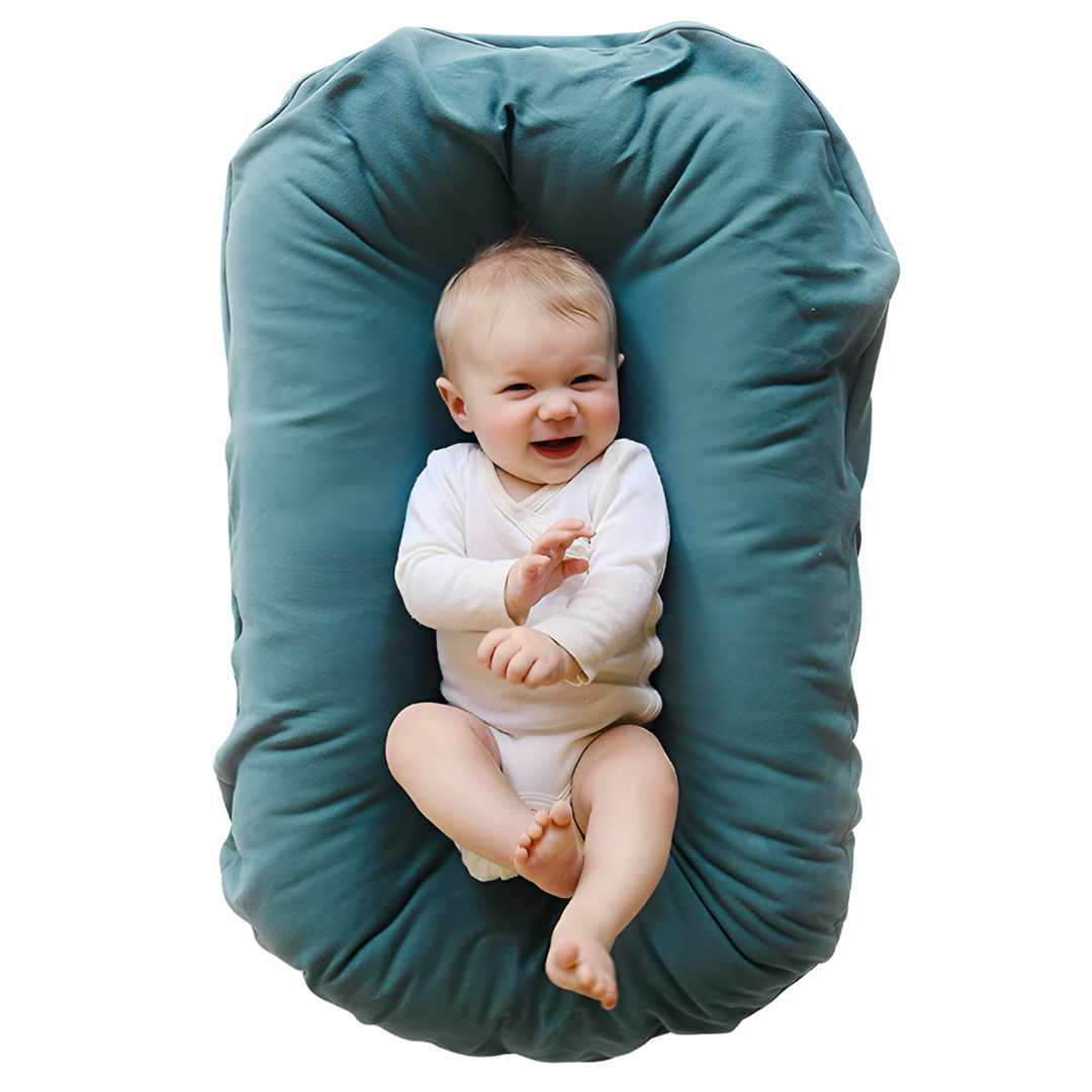Laughing baby in a white bodysuit lies in a teal nest cushion, seen from above 2