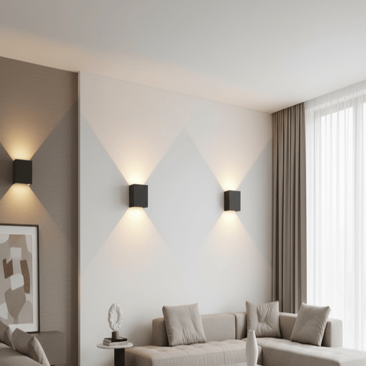 Three black square wall lights on a white living room wall, each casting upward and downward cones of warm light beside a sofa.