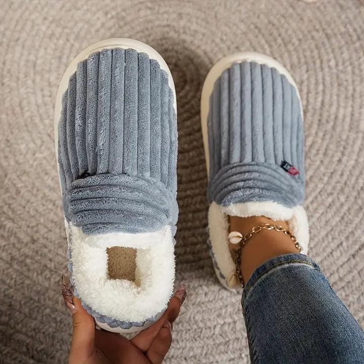 Top view of gray-blue ribbed slippers on a woven rug, one held in a hand and one worn with a shell anklet.