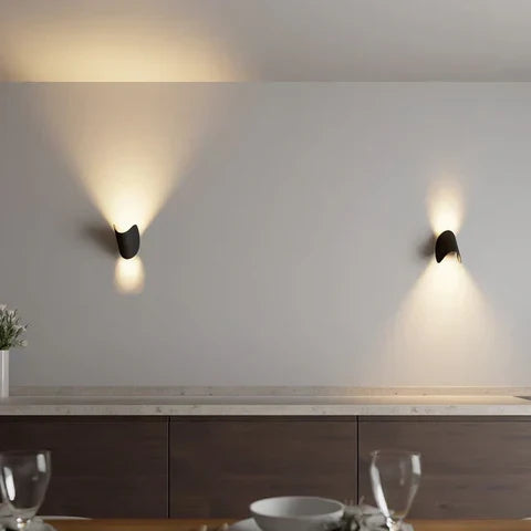 Two small black metal wall lights on a pale wall above a dining table, each throwing warm light up and down.