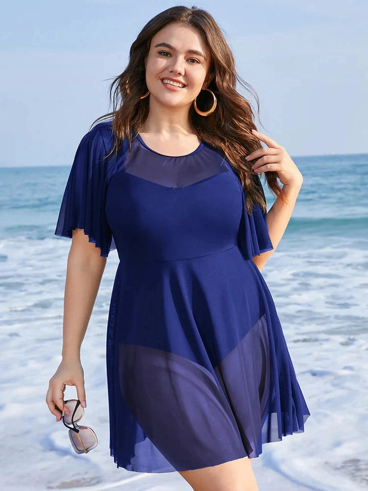 Frau im blauen Strandkleid am Meer, lächelnd, mit Sonnenbrille in der Hand.