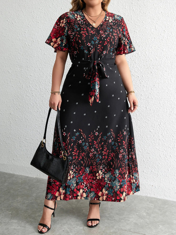 Frau in langem, schwarzem Blumenkleid mit Gürtel, hält schwarze Handtasche, trägt Sandalen.