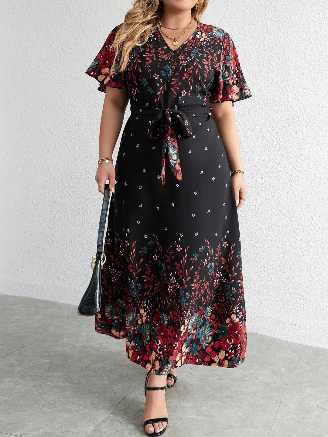Frau in langem, schwarzem Blumenkleid mit Gürtel, V-Ausschnitt, kurzen Ärmeln, Sandalen, Handtasche.