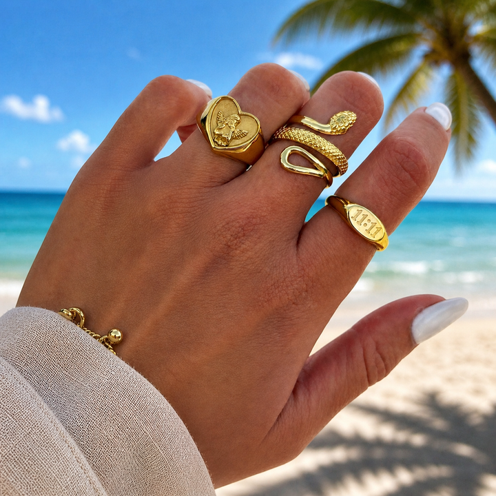 Hand wearing the gold snake ring on the middle finger alongside a heart signet ring and an oval ring, on a sunny beach. 4