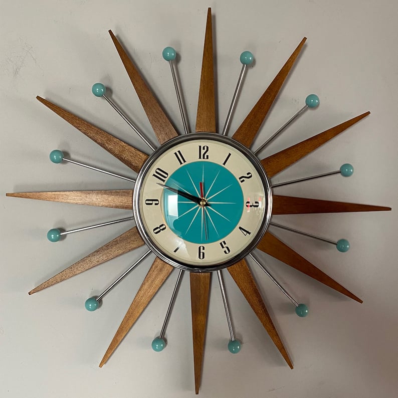 Starburst wall clock detail on a gray wall, showing a chrome-rimmed cream and teal dial with black numerals and a red second hand.