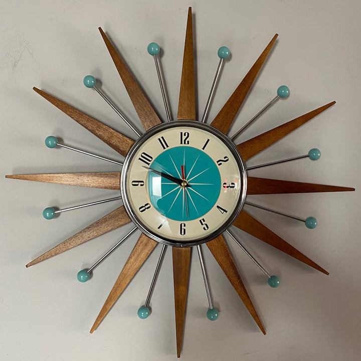 Starburst wall clock detail on a gray wall, showing a chrome-rimmed cream and teal dial with black numerals and a red second hand.