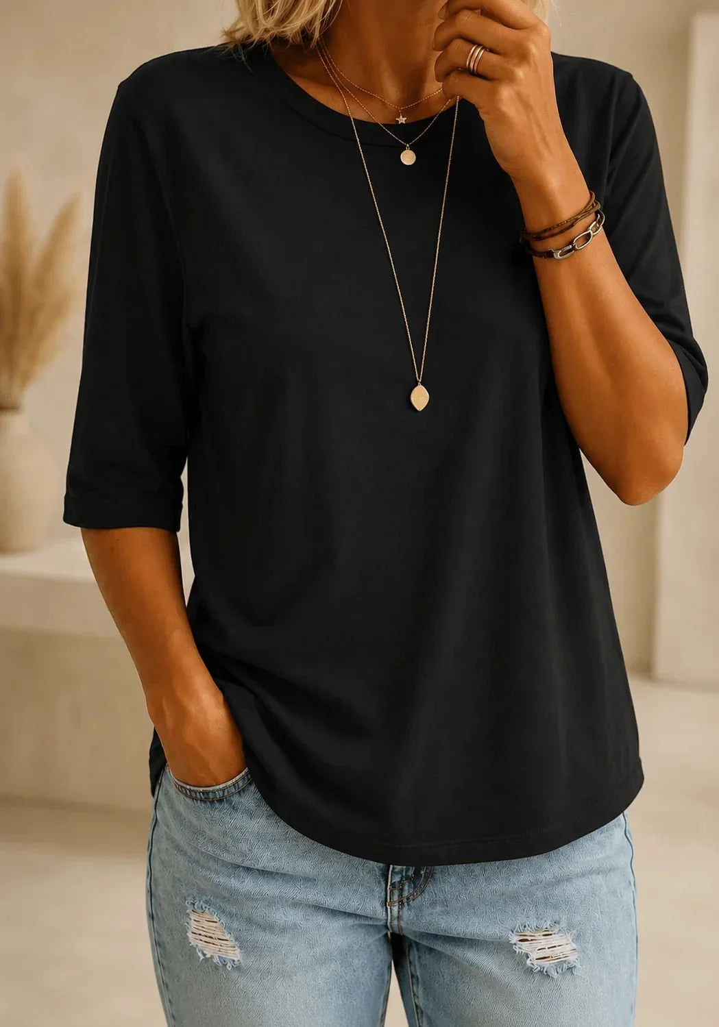 Woman wearing a black half-sleeve round neck tee with layered gold necklaces and light ripped jeans, hand resting in her pocket. 1