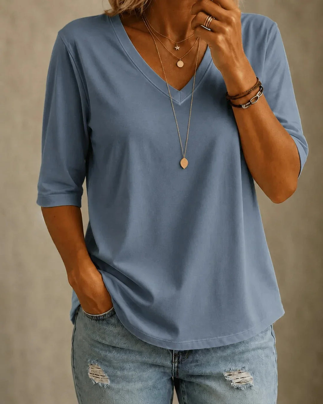 Woman wearing a slate blue half-sleeve V-neck tee with layered gold necklaces and distressed light-wash jeans, front view, one hand in pocket. 1