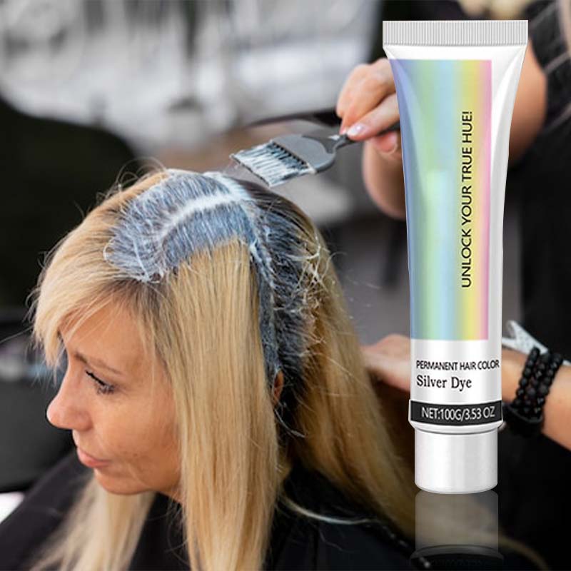 Stylist brushing silver color cream onto a blonde woman's roots in a salon, with the tube shown at right. 4