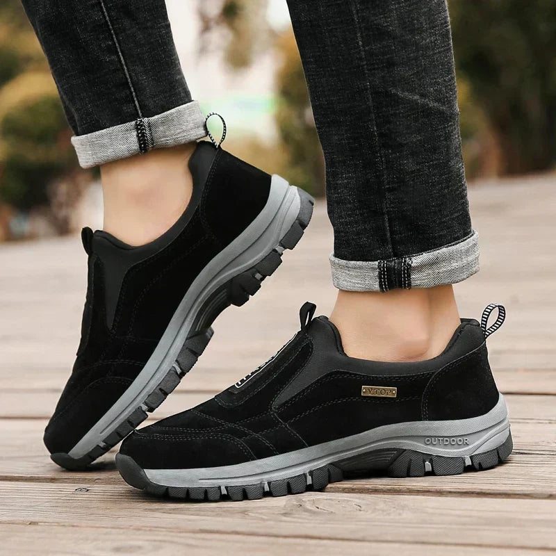 Black slip-on shoes worn on a wooden deck with rolled dark jeans, showing the heel loop and thick gray outsole. 4