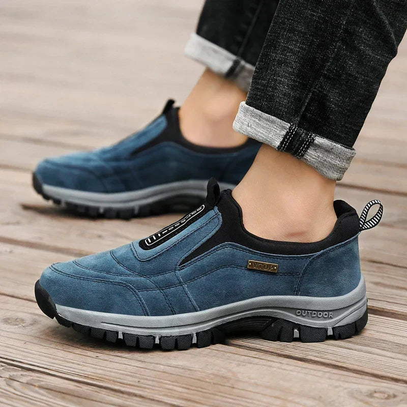 Blue slip-on shoes worn by a model in rolled dark jeans on weathered wood planks, showing the black collar and gray lugged sole. 5