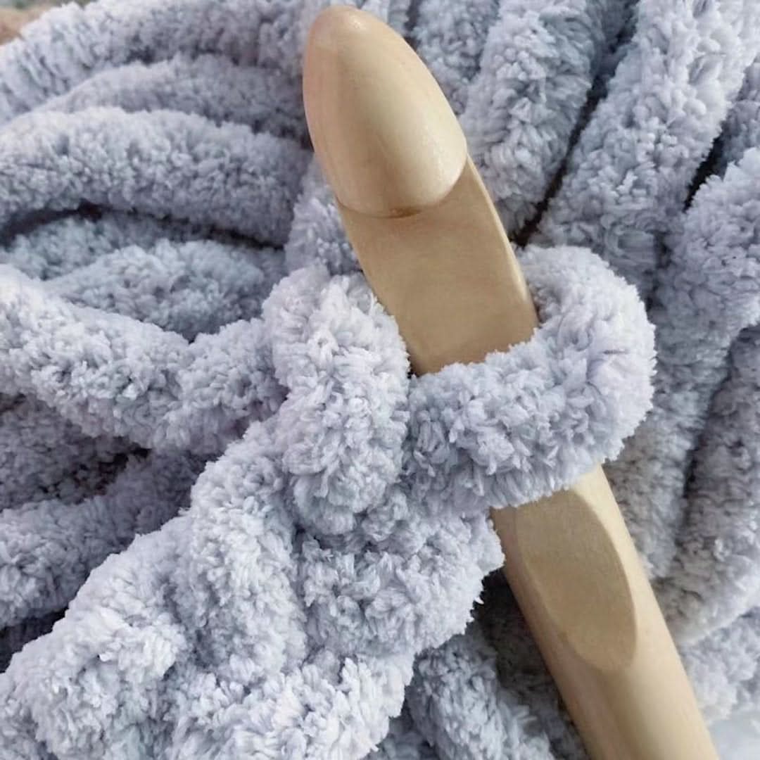 detail of a large wooden crochet hook looped through soft, fluffy light gray-blue chunky yarn stitches.