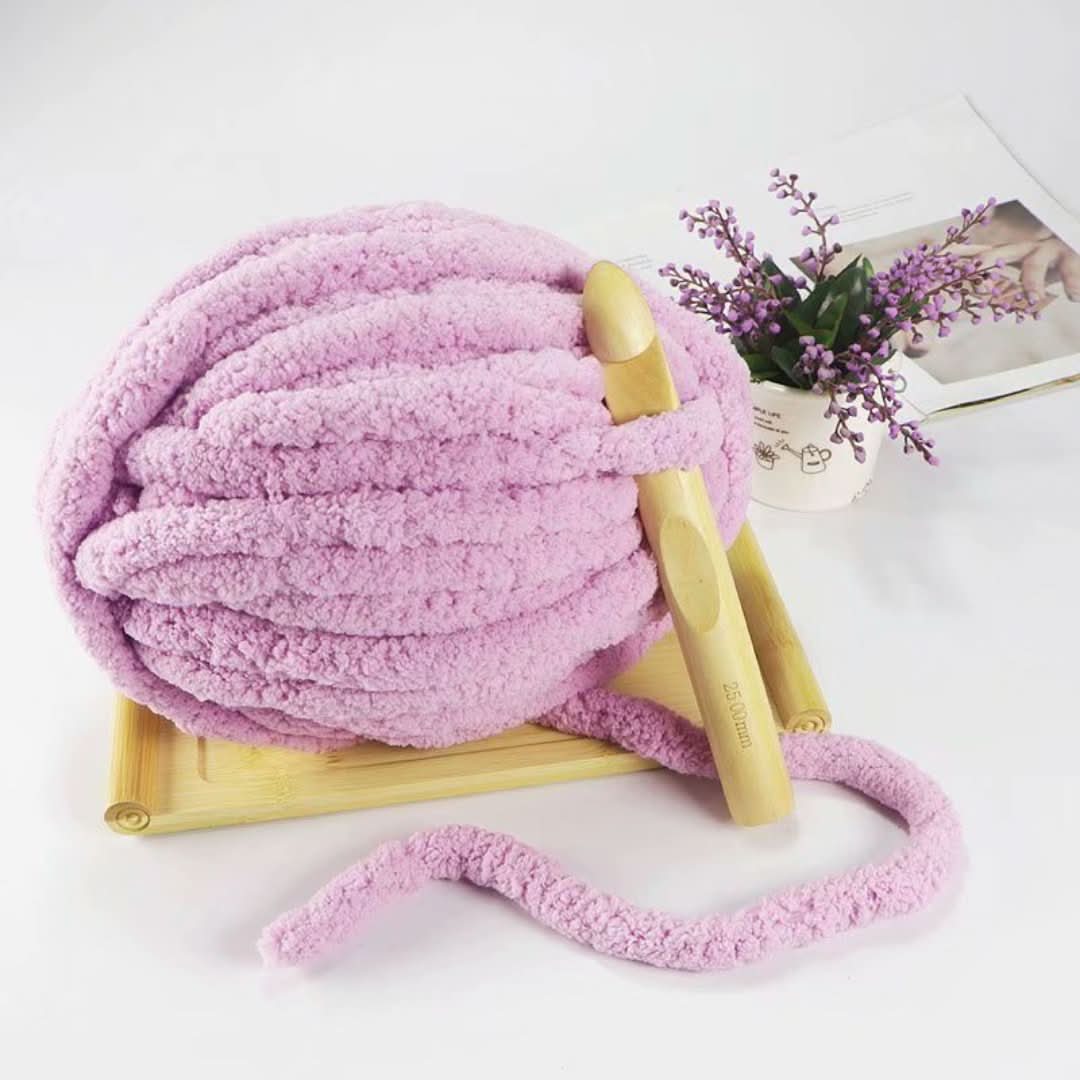 Pink skein of chunky yarn with a large wooden crochet hook on a bamboo stand beside a potted flower.