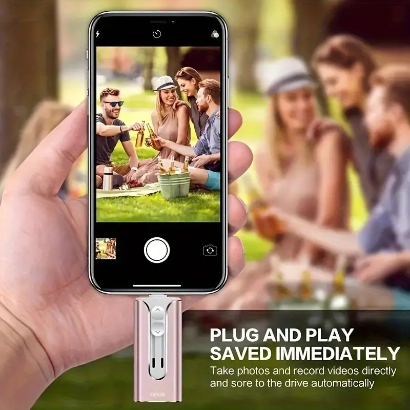 Hand holding a phone camera screen showing friends at a picnic, with a pink drive plugged into the phone's bottom port.