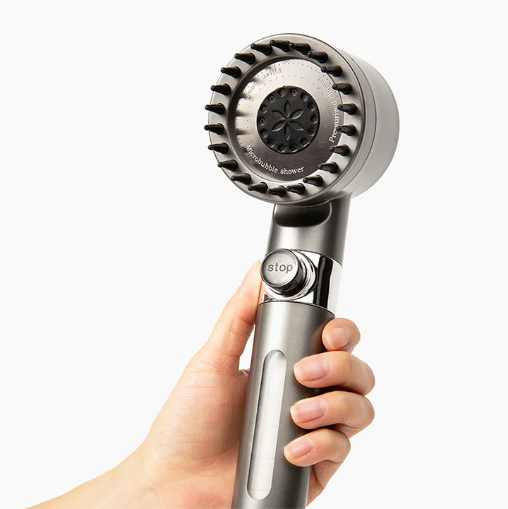 Hand holding the grey shower head upright, showing the bristled spray face, the round stop button and the clear filter window.