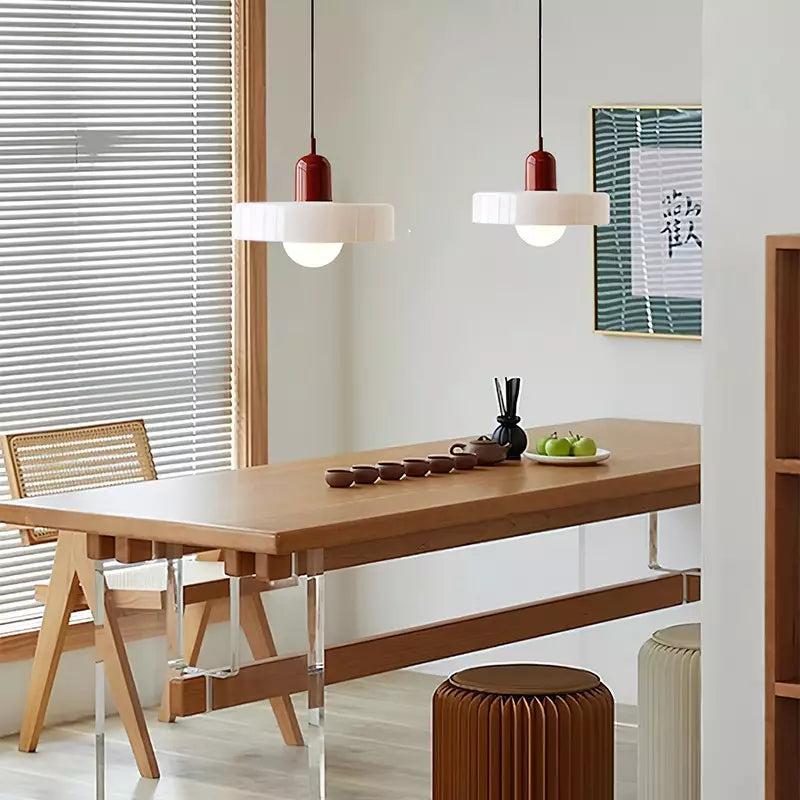 Two white glass pendants with red caps hang above a long wooden table by a window with blinds 36
