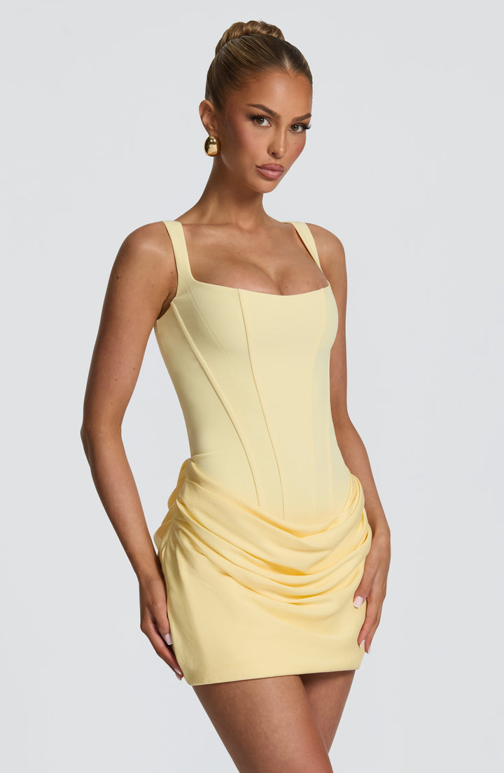 Angled view of the pale yellow mini dress, highlighting the curved corset seams and the ruched, draped hip detail. 27