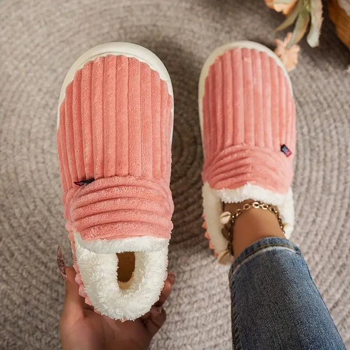 Top view of coral pink ribbed slippers on a woven rug, one held in a hand and one worn on a foot.