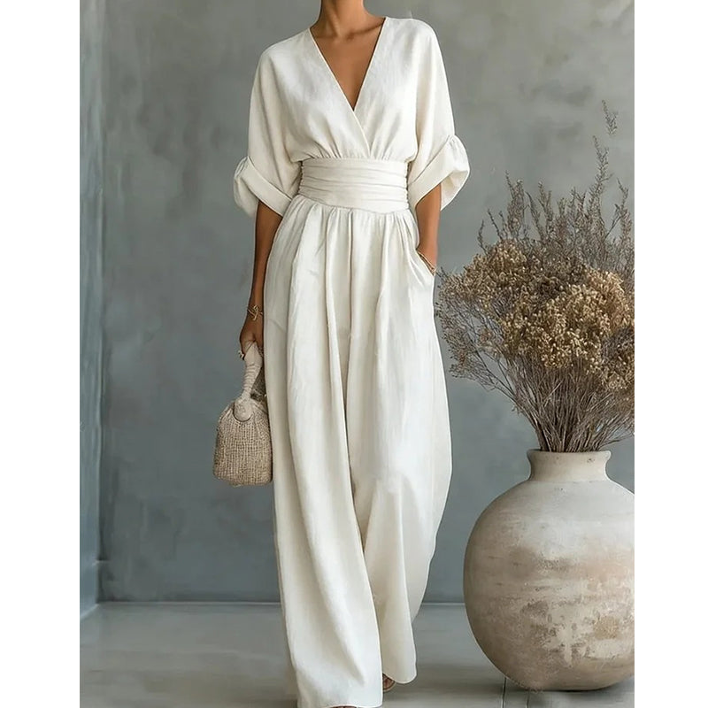 Woman in a cream white wide-leg jumpsuit with a wrap V-neck and cinched waist, holding a woven bag beside a large vase. 1