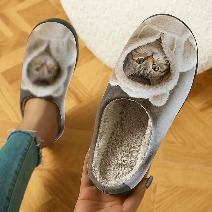 Top-down view of one slipper held in a hand, showing the upside-down cat print and soft fleece lining, with the other slipper worn. 2
