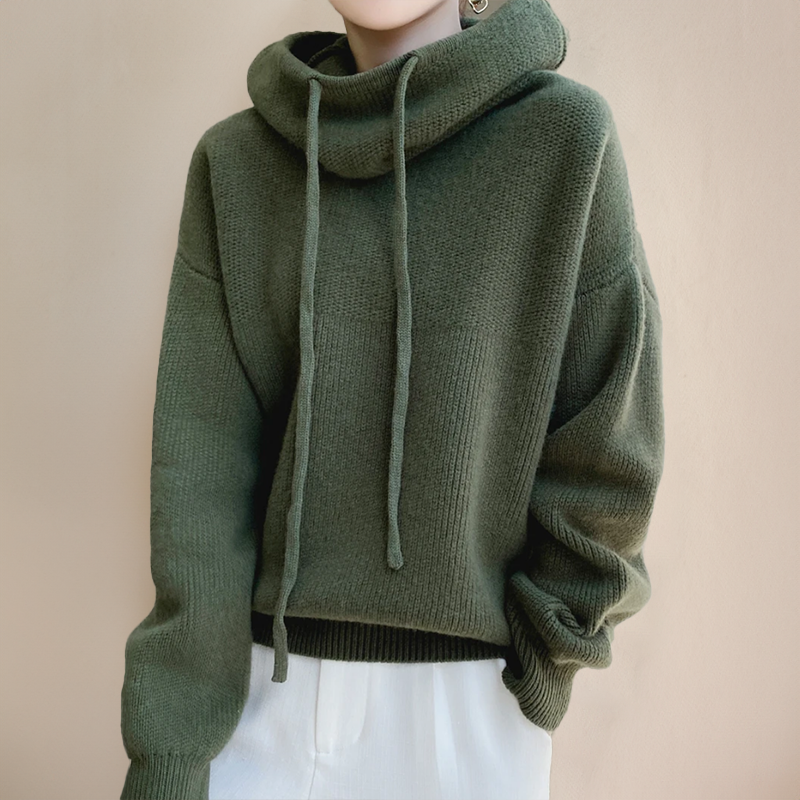 Dark olive green drawstring cowl-neck sweater with dropped shoulders, worn with white trousers against a beige wall 3
