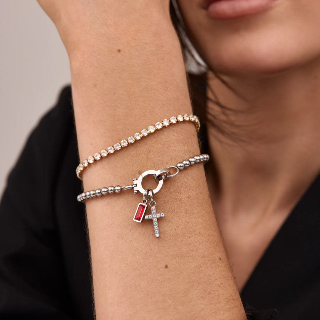Silver-tone bead bracelet worn on a wrist with a red baguette stone charm and crystal cross charm.