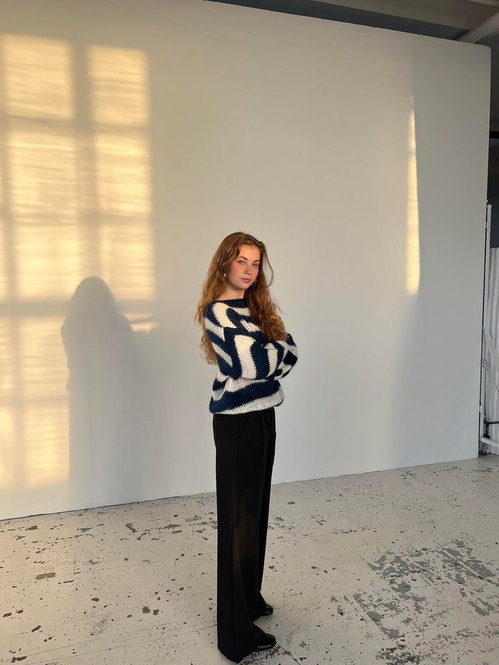 Frau in gestreiftem Pullover, schwarze Hose, steht vor weißer Wand, Sonnenlicht im Hintergrund.