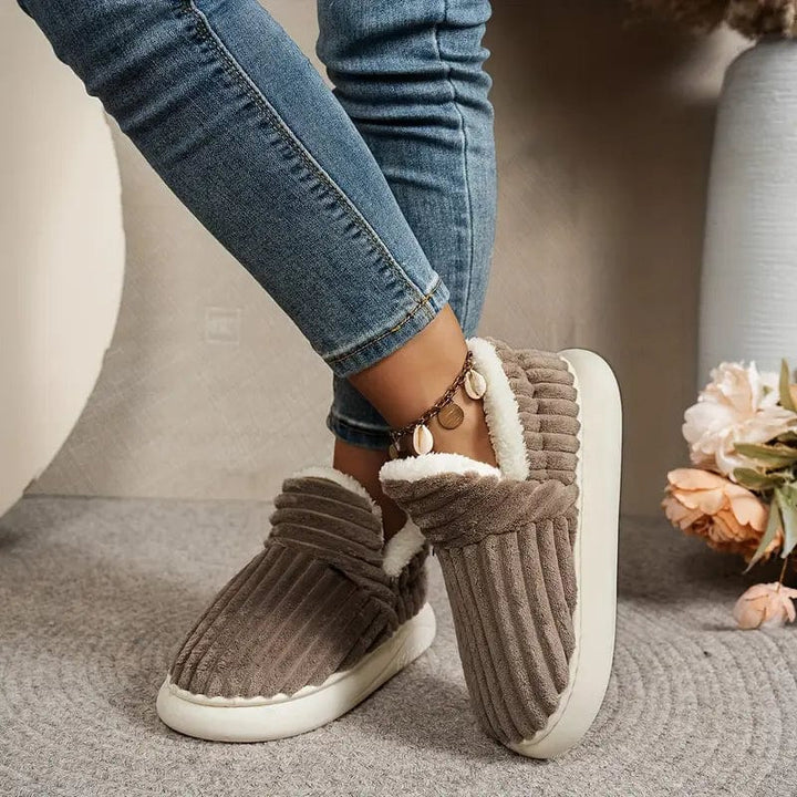 Feet in brown ribbed fleece-lined slippers and jeans, crossed one over the other on a gray rug near flowers.