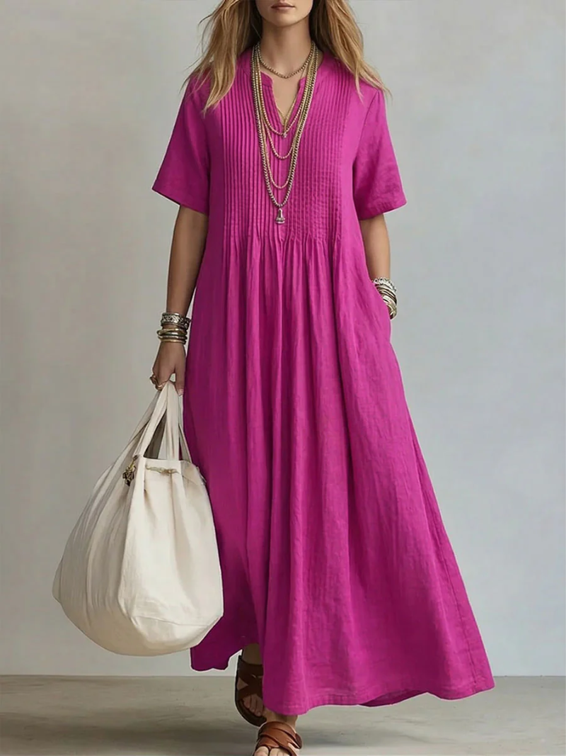 Model wears the short-sleeve pleated dress in bright magenta, carrying a cream tote and wearing brown sandals.