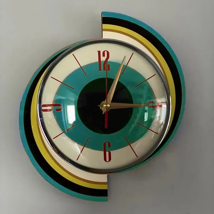 Swirl wall clock front view on a gray wall, with offset teal, black, yellow and cream arcs behind a teal-ringed dial.