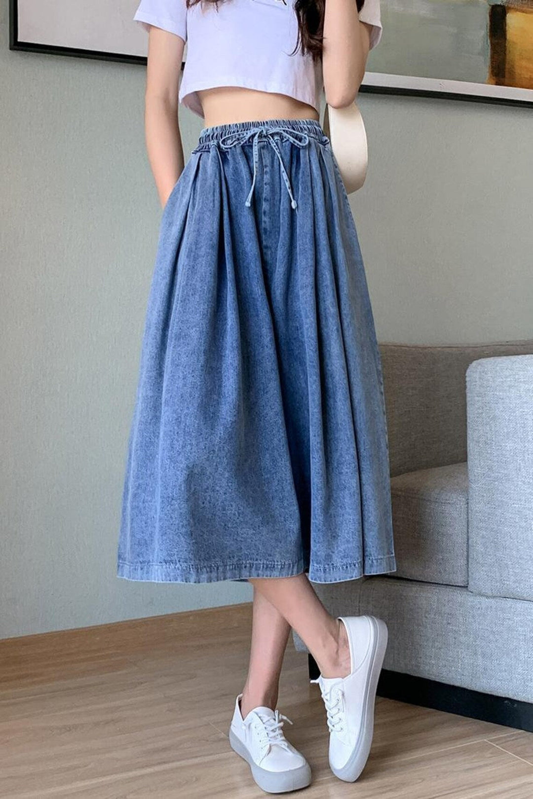 Frau trägt blauen Jeansrock mit Kordelzug, weißes T-Shirt und weiße Sneaker in modernem Wohnzimmer.