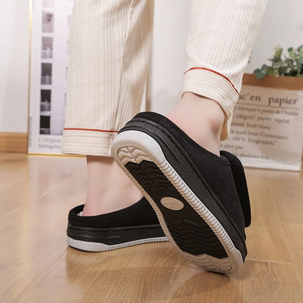 Rear view of a person in cream striped pants wearing black slippers, with one foot raised to show the treaded non-slip sole. 8