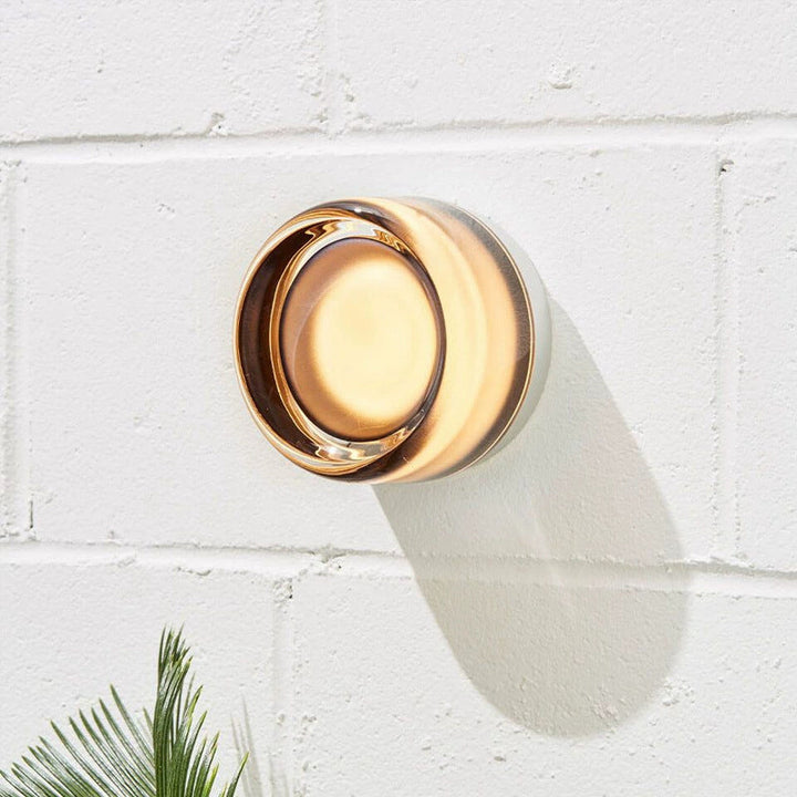 Glowing amber glass sconce with a dimpled center and white base, mounted on a white brick wall beside a palm frond. 1