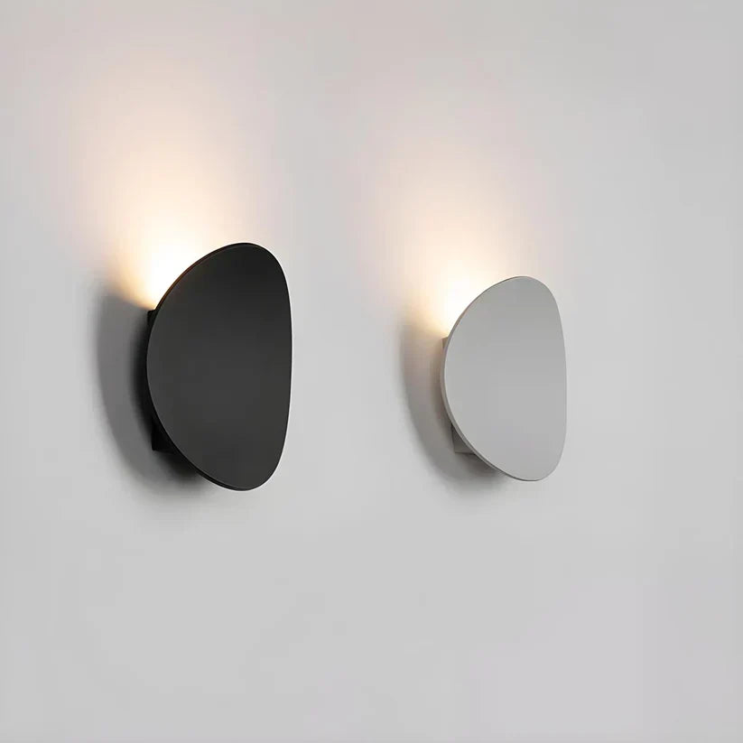 Black and white versions of the oval wall lamp side by side on a plain wall, both glowing upward. 5
