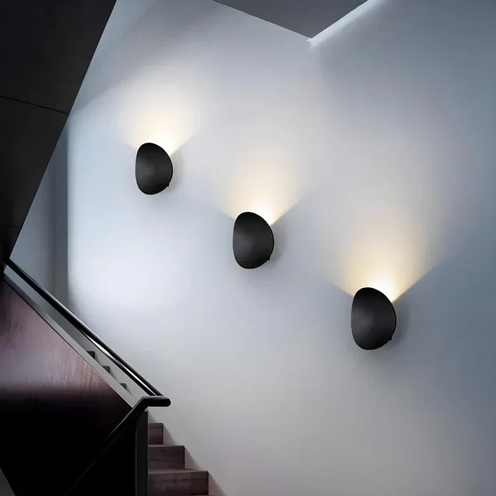 Three black oval wall lamps staggered up a stairwell wall, each throwing warm light upward. 7