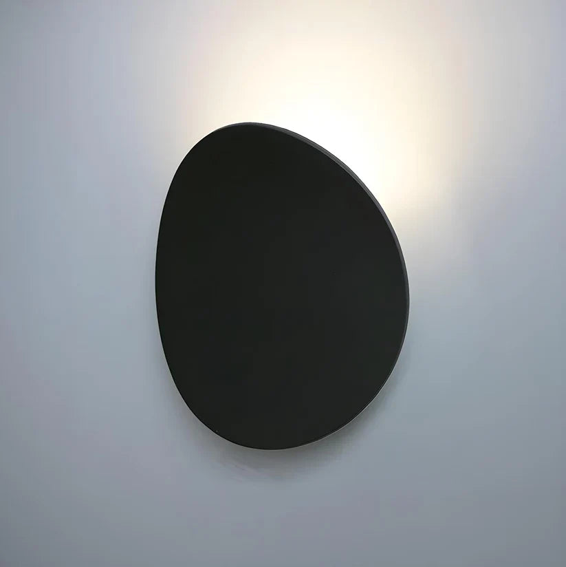 Front view of the matte black oval wall lamp on a pale wall with a soft halo of light behind its top edge. 8