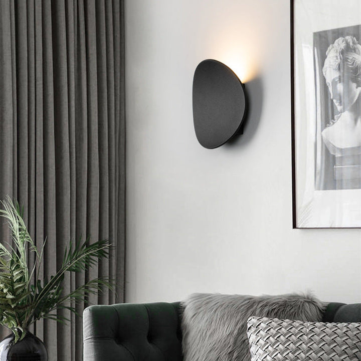 Single black disk wall lamp lit above a dark green sofa, beside gray curtains and a framed statue print. 3