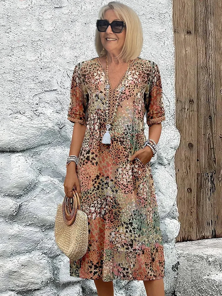 Model wearing the V-neck midi dress in an earthy multicolor speckled print with peach, olive and brown tones, holding a straw bag. 5