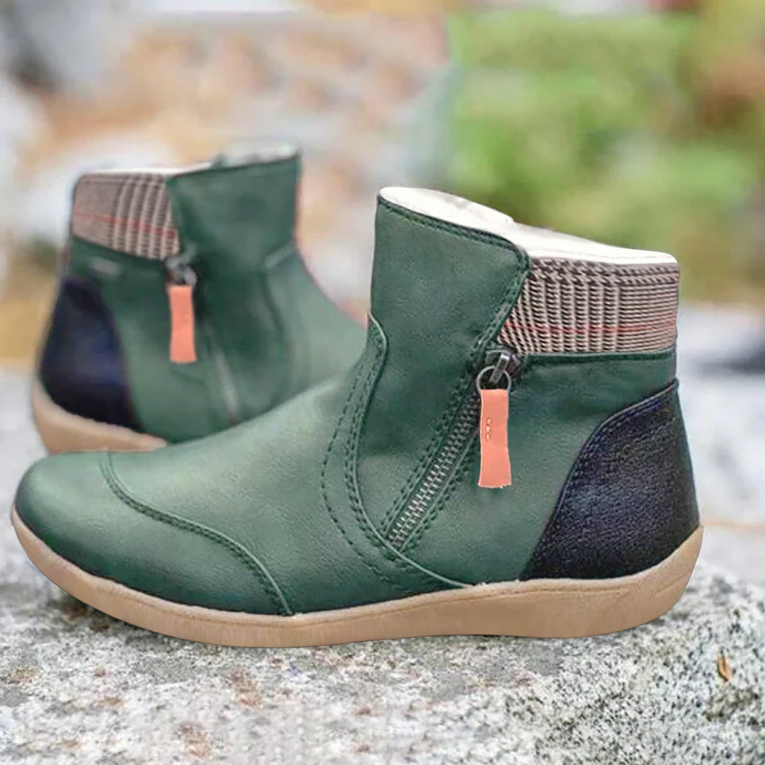 Pair of green ankle boots with side zipper, orange pull tab, plaid collar and black heel panel, standing on a rock outdoors.